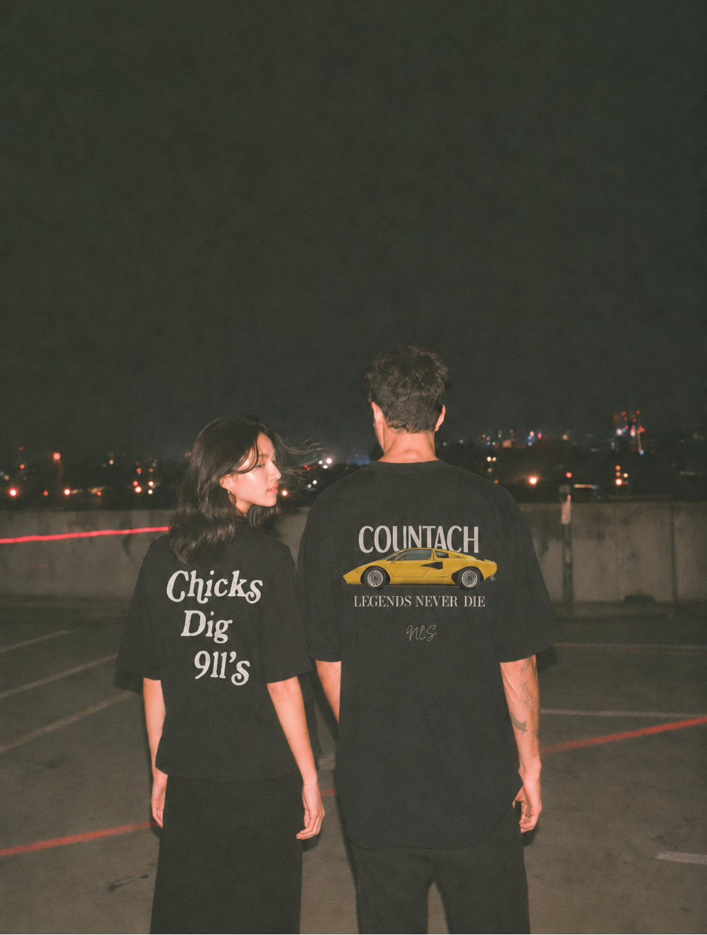 Two people standing on a rooftop at night, seen from behind, wearing matching black graphic t-shirts. The shirt on the left reads 'Chicks Dig 911's' in white text, and the shirt on the right reads 'COUNTACH LEGENDS NEVER DIE' with a yellow car graphic.